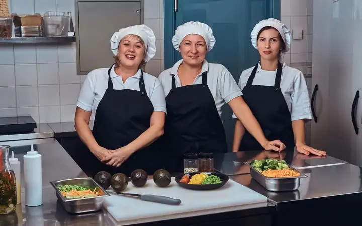Tres cocineras con filipina y mandil junto a ingredientes frescos en una cocina de acero inoxidable
